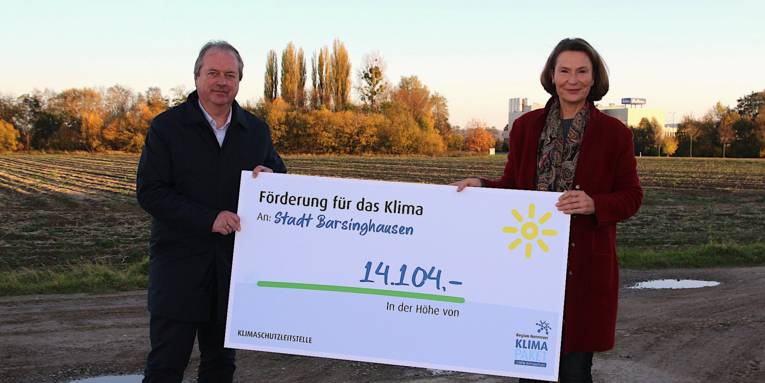 Ein Mann und eine Frau halten einen symbolischen Scheck über 14.104 Euro. Sie stehen auf einer Feldwegkreuzung